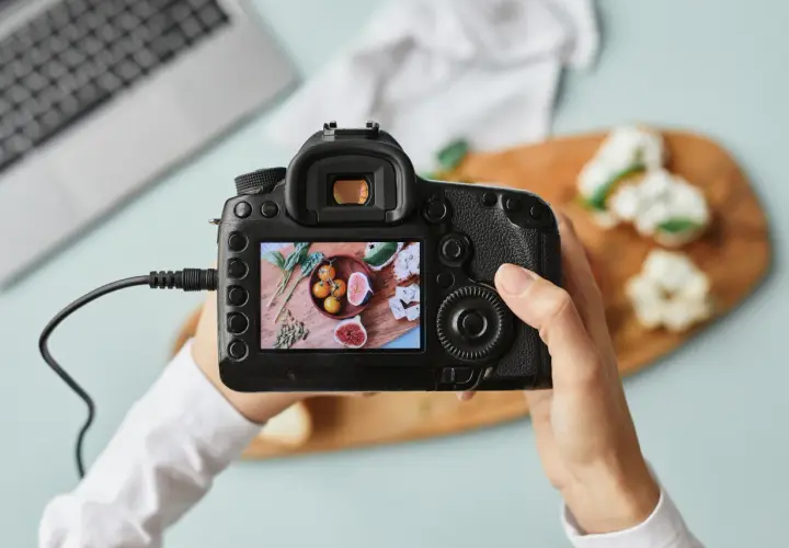 Dłonie trzymające aparat fotograficzny, na którego ekranie widać zdjęcie jedzenia.