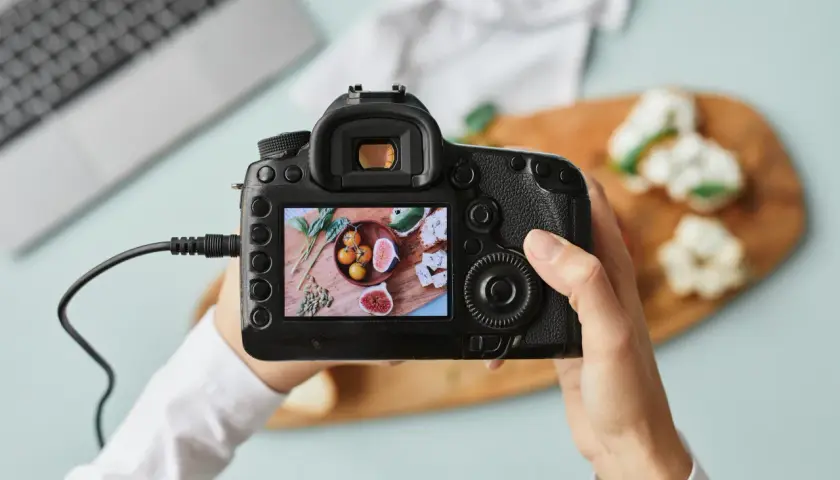 Dłonie trzymające aparat fotograficzny, na którego ekranie widać zdjęcie jedzenia.