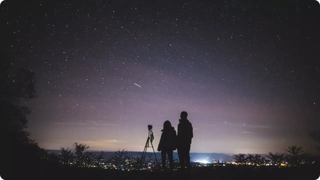 Para zrobić zdjęcie gwiazd iPhonem, ustaw aparat na statywie i skieruj go na nocne niebo. Dwie sylwetki obserwują gwiazdy, jedno z nich przygotowuje się do fotografowania.