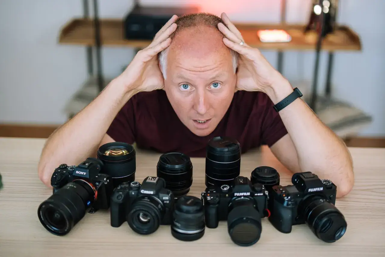 Zastanawiasz się, jakie aparaty fotograficzne wybrać? Na stole leży kilka modeli Sony, Canon i Fujifilm, a mężczyzna z rękami na głowie wygląda na zamyślonego.